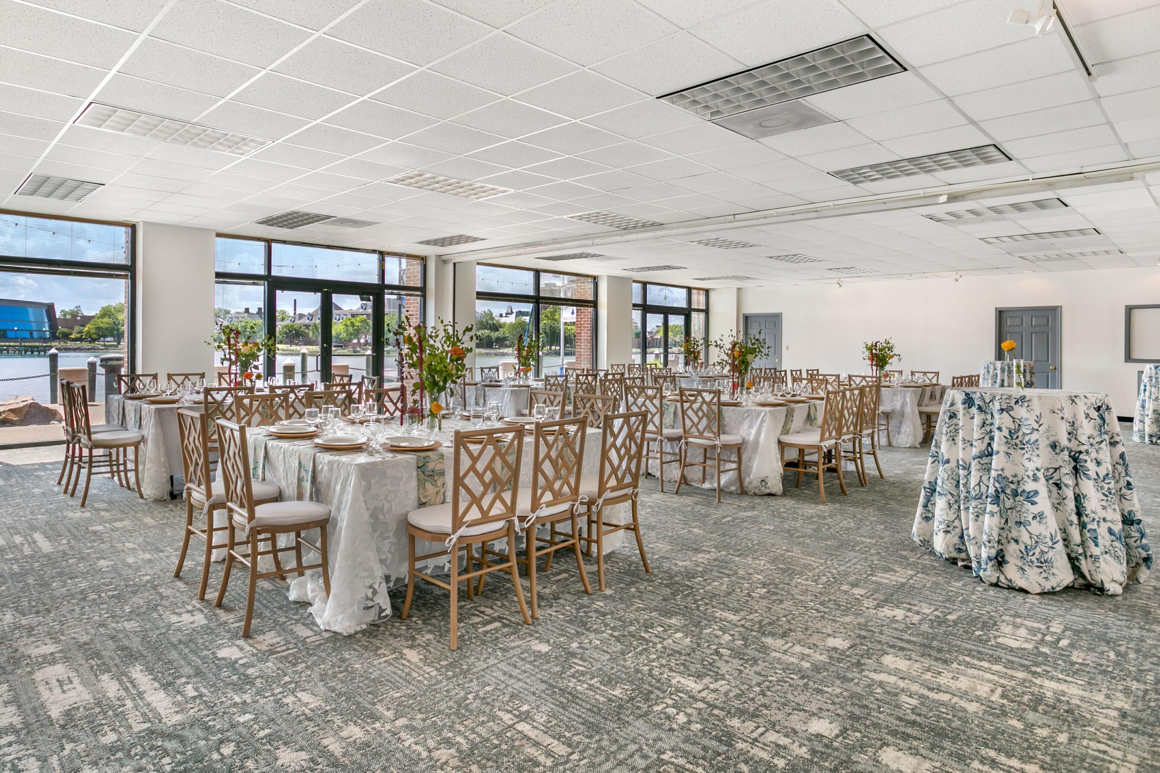 Un amplio salón de banquetes con varias mesas redondas dispuestas para comer, grandes ventanales que ofrecen vistas al exterior y una alfombra estampada cubriendo el suelo.