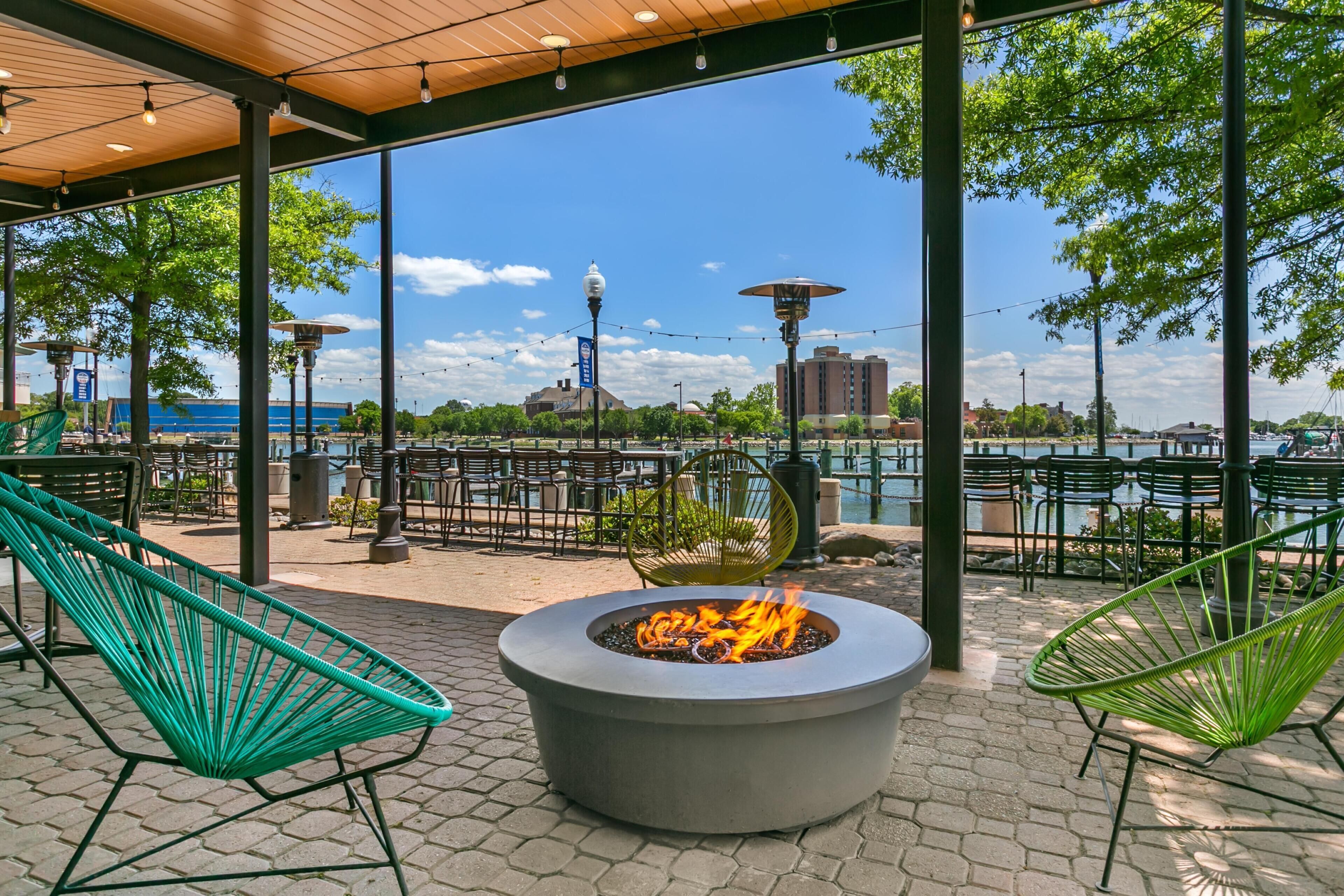 An outdoor seating area with a fire feature, chairs, and a view of a body of water and buildings.