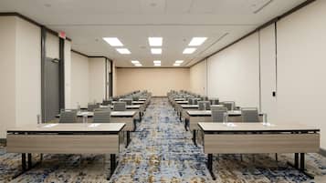 Une salle de conférence avec des rangées de tables et de chaises, un sol recouvert de moquette et des luminaires au plafond.