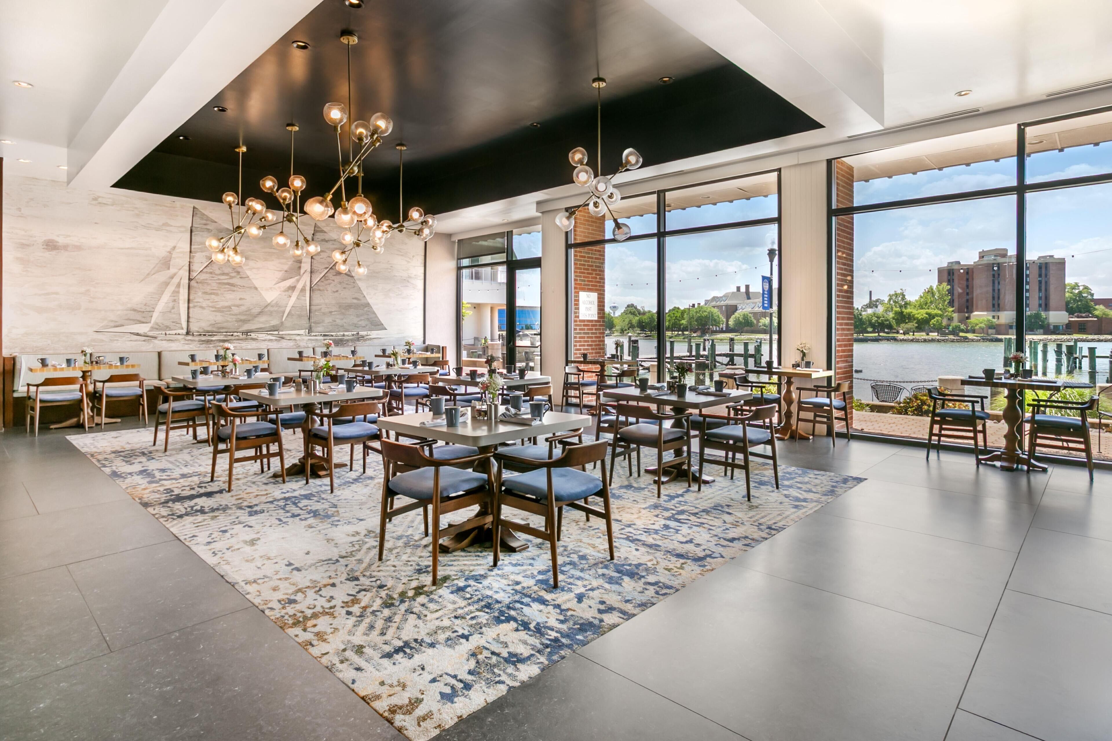A spacious dining area with multiple tables and chairs, large windows offering views of a river and buildings, and a modern chandelier.