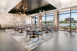 A spacious dining area with multiple tables and chairs, large windows offering views of a river and buildings, and a modern chandelier. - The Landing at Hampton Marina, Tapestry Collection by Hilton (Hampton)