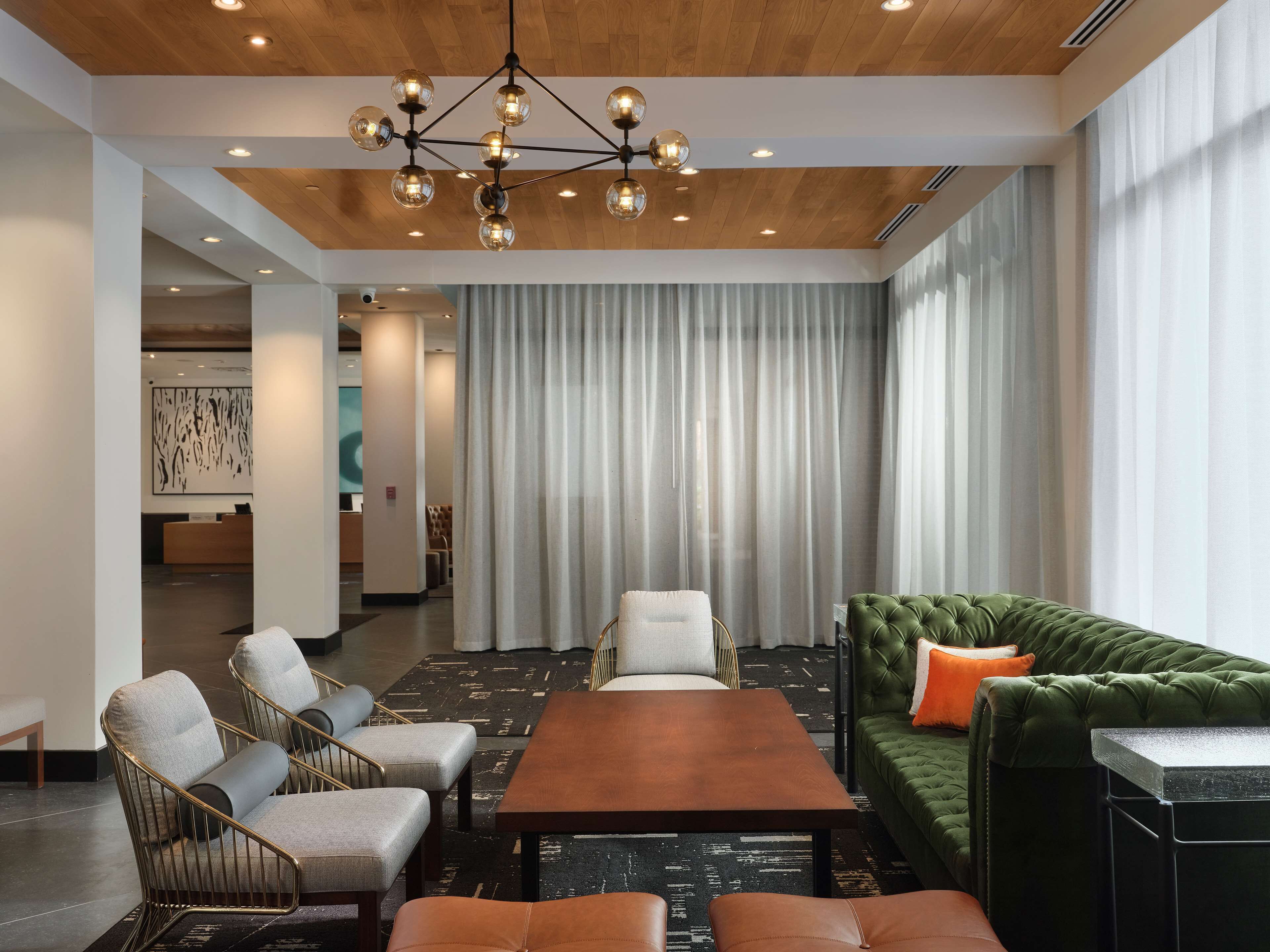 Un hall d’hôtel moderne avec un plafond en bois, un lustre central, un canapé vert et un comptoir d’accueil.