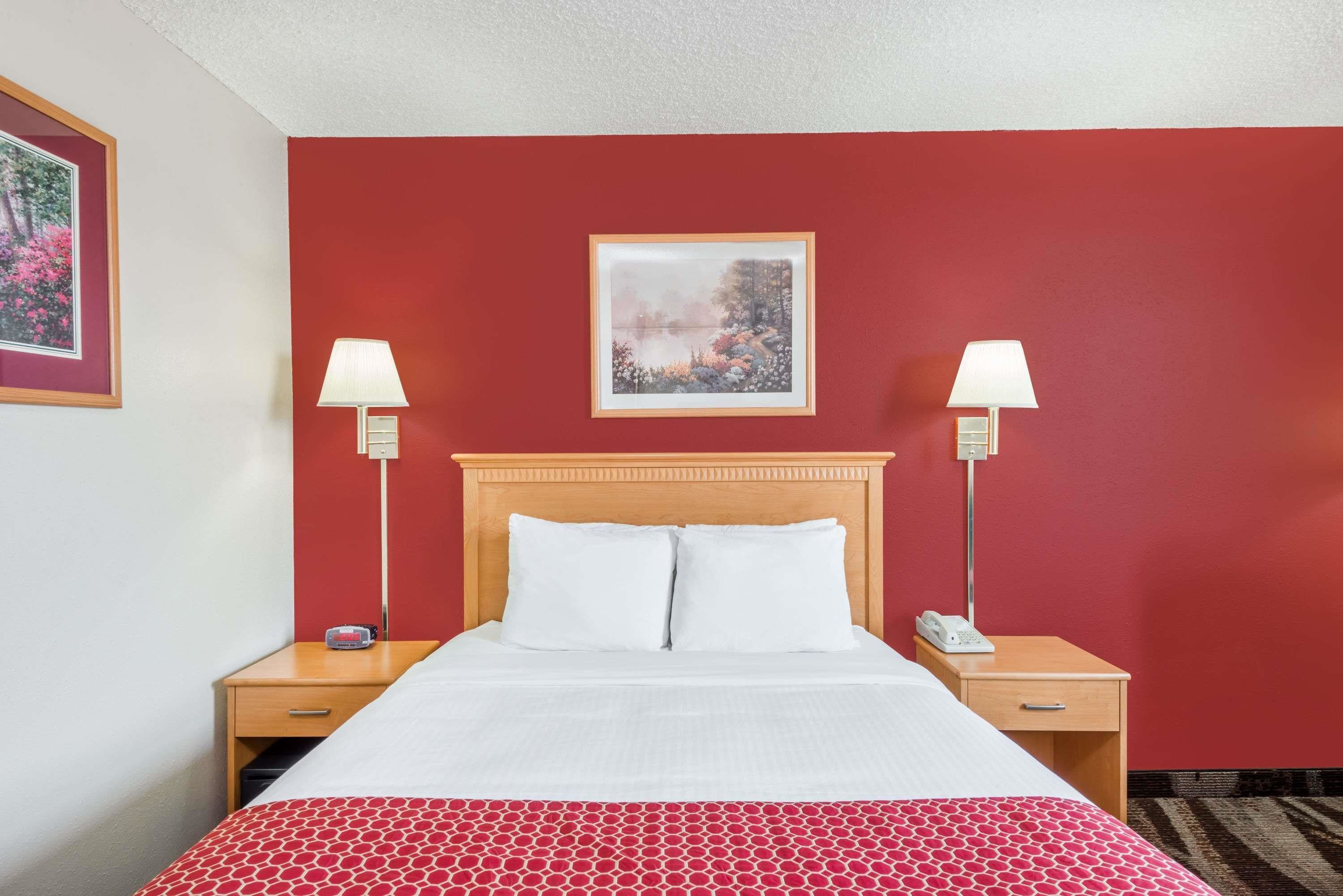 A hotel room with a red wall, a wooden headboard, two bedside tables with lamps, a bed with white bedding, and a framed picture on the wall.