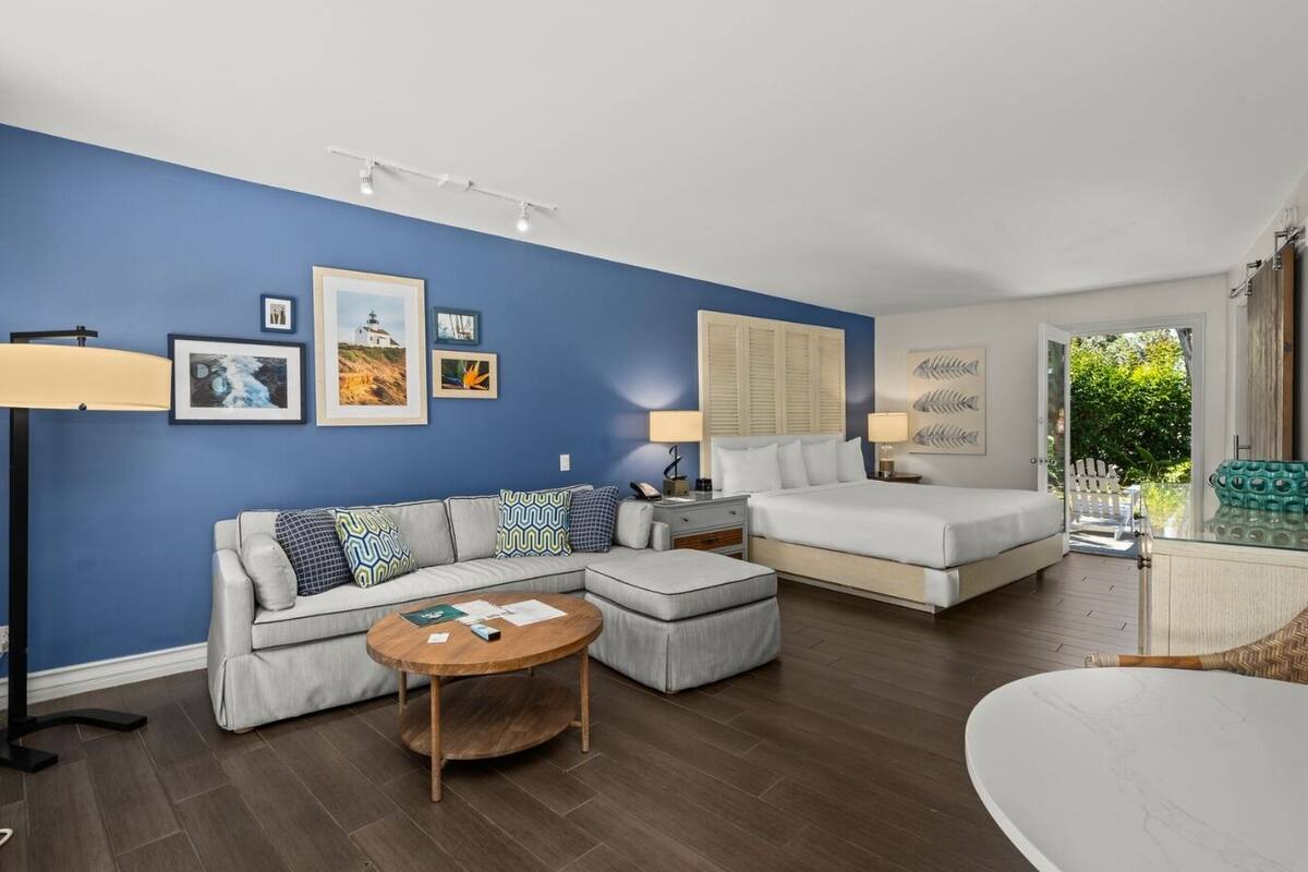 A modern living room with a sofa, ottoman, and coffee table, a bed with a nightstand, and a dining area.