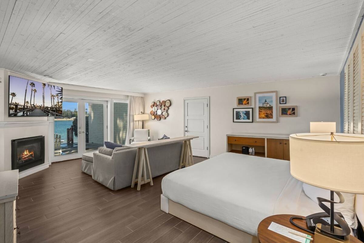 A modern hotel room with a large bed, a desk, a fireplace, and a balcony with a view of palm trees and a beach.