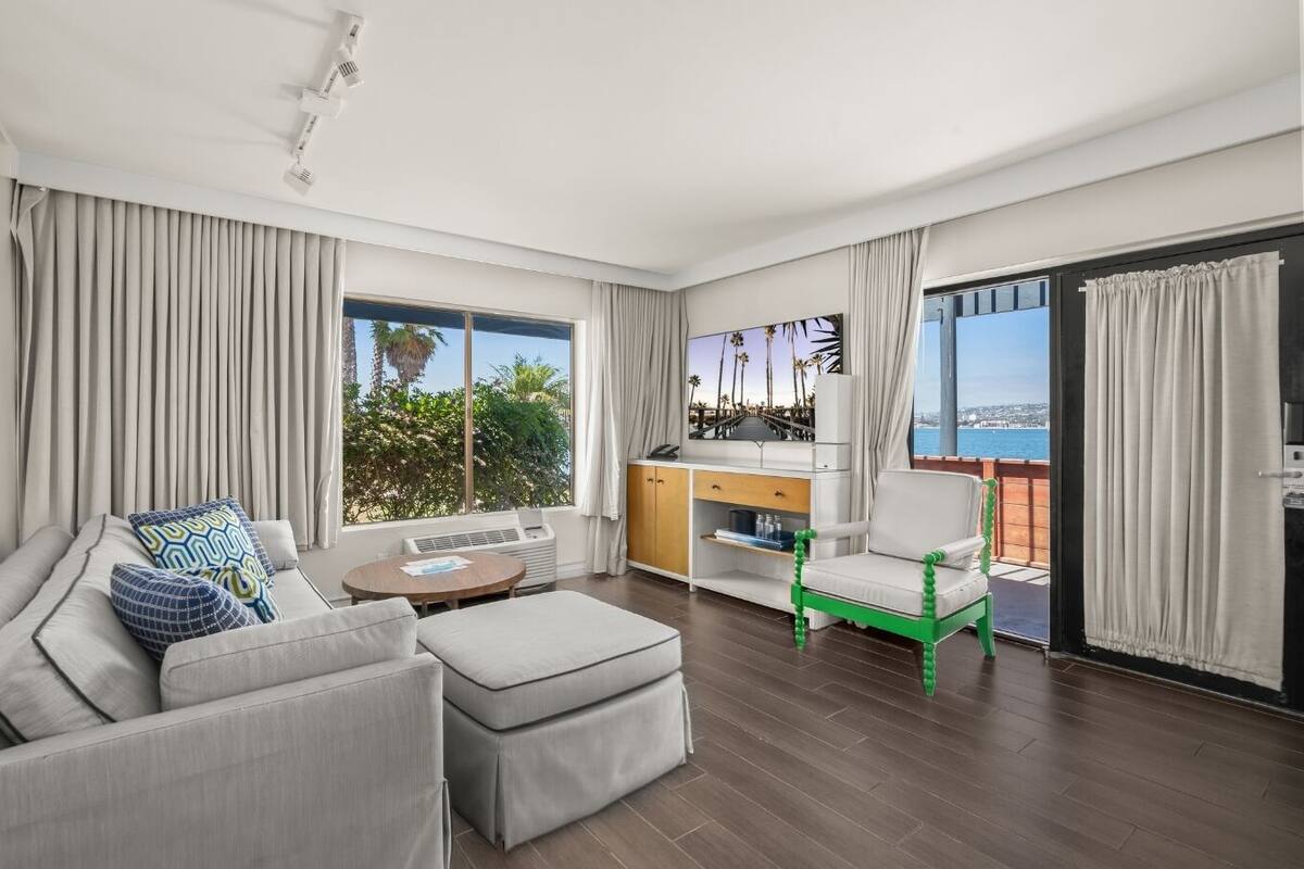 A modern living room with a sofa, a coffee table, and a green chair. There is a TV and a small table with a laptop. The room has large windows with curtains and a view of the sea.