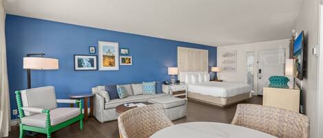 A modern hotel room with a blue accent wall, a sofa, a bed, a dining table, and a TV.
