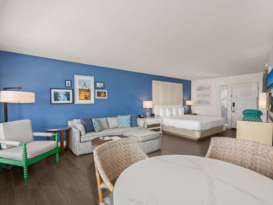 A modern hotel room with a blue accent wall, a sofa, a bed, a dining table, and a TV.