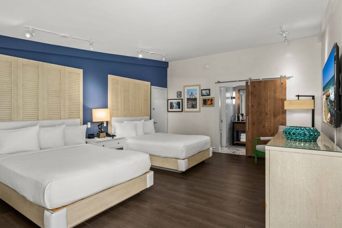 A hotel room with two beds, a wooden wardrobe, a desk with a lamp, and a painting on the wall.