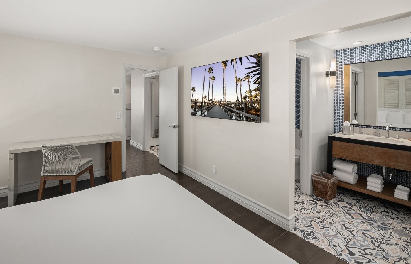 A hotel room with a bed, a desk, a chair, a bathroom with a sink and mirror, and a large picture on the wall.