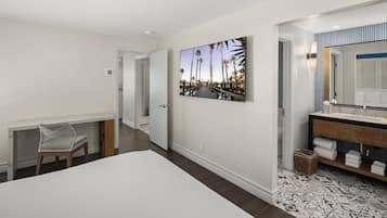 A hotel room with a bed, a desk, a chair, a bathroom with a sink and mirror, and a large picture on the wall.