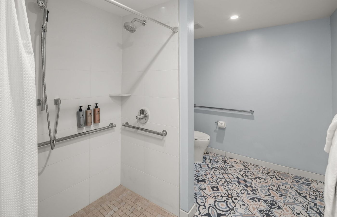 Un baño moderno con azulejos blancos, una ducha con cabezal de ducha manual, un inodoro con barra de apoyo y un suelo estampado.