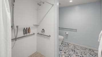Un baño moderno con azulejos blancos, una ducha con cabezal de ducha manual, un inodoro con barra de apoyo y un suelo estampado.