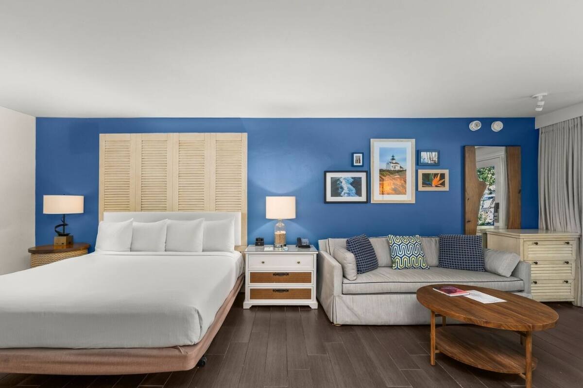 A hotel room with a bed, a sofa, a coffee table, and a dresser. There are wall-mounted lamps, a nightstand, and framed pictures on the wall.