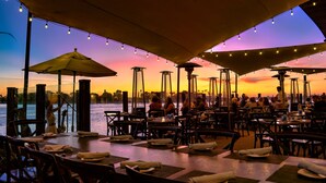 An outdoor dining area with tables and chairs, umbrellas, and string lights against a sunset backdrop.