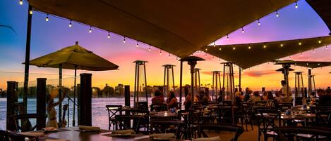 An outdoor dining area with tables and chairs, umbrellas, and string lights against a sunset backdrop.