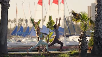 Dos personas practicando yoga en un espacio al aire libre, con barcos y banderas al fondo.