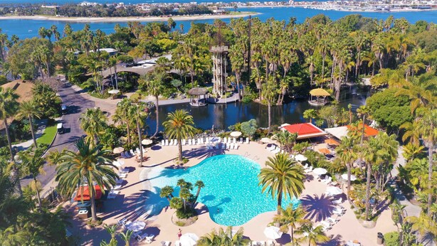 An aerial view of a resort with a central swimming pool, palm trees, and a view of the ocean and beach.