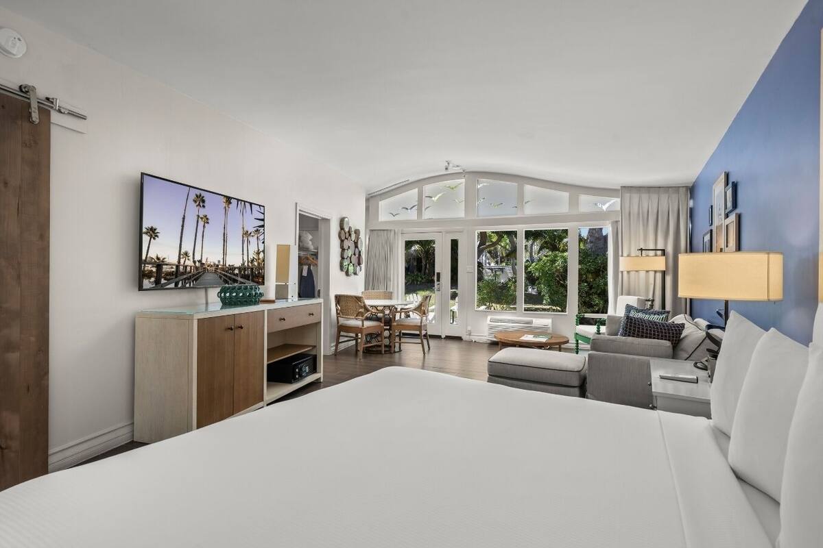 A hotel room with a large bed, a desk, a chair, and a view of greenery through large windows.