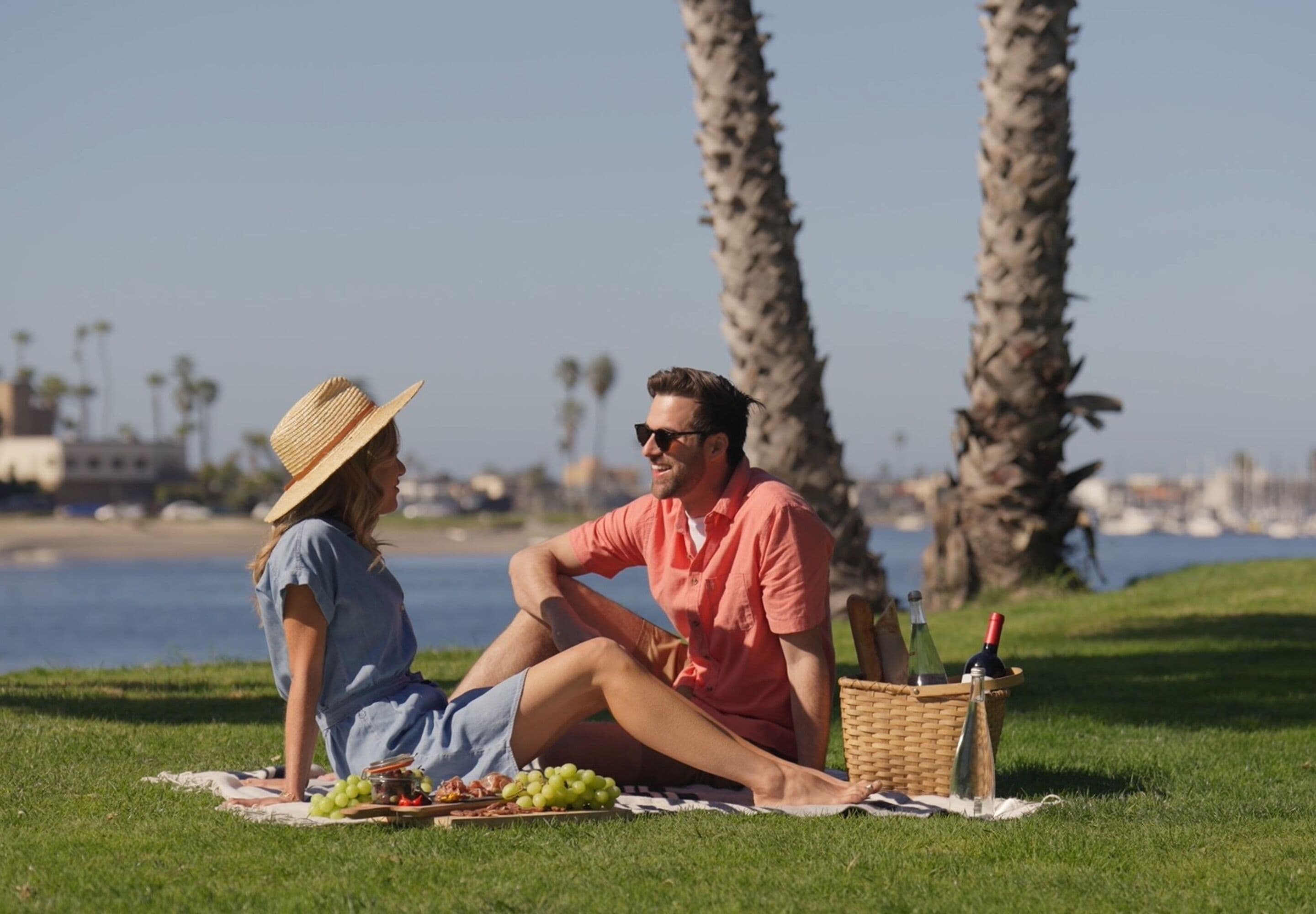 Una pareja sentada sobre una manta con una preparación para picnic, incluyendo uvas y una botella, en un parque con palmeras y un cuerpo de agua al fondo.