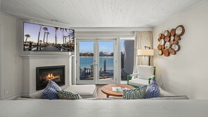 A living room with a fireplace, a television, a sofa, a chair, a round coffee table, and a view of the ocean through glass doors.