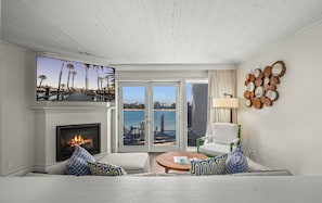 A living room with a fireplace, a television, a sofa, a chair, a round coffee table, and a view of the ocean through glass doors.