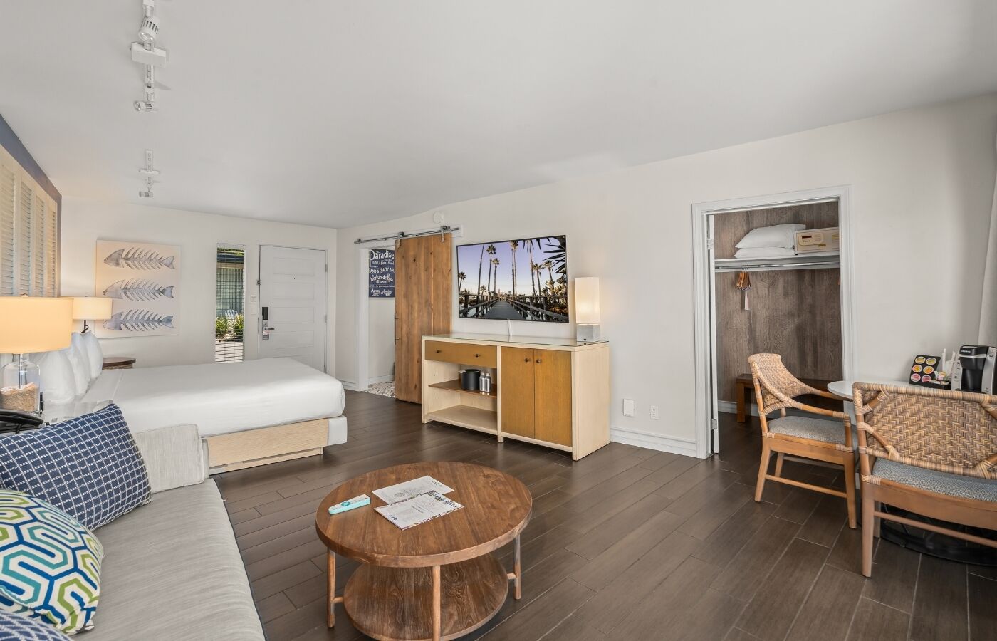 A modern hotel room with a bed, a wooden desk, a round coffee table, a sofa, and a dining area with wicker chairs.