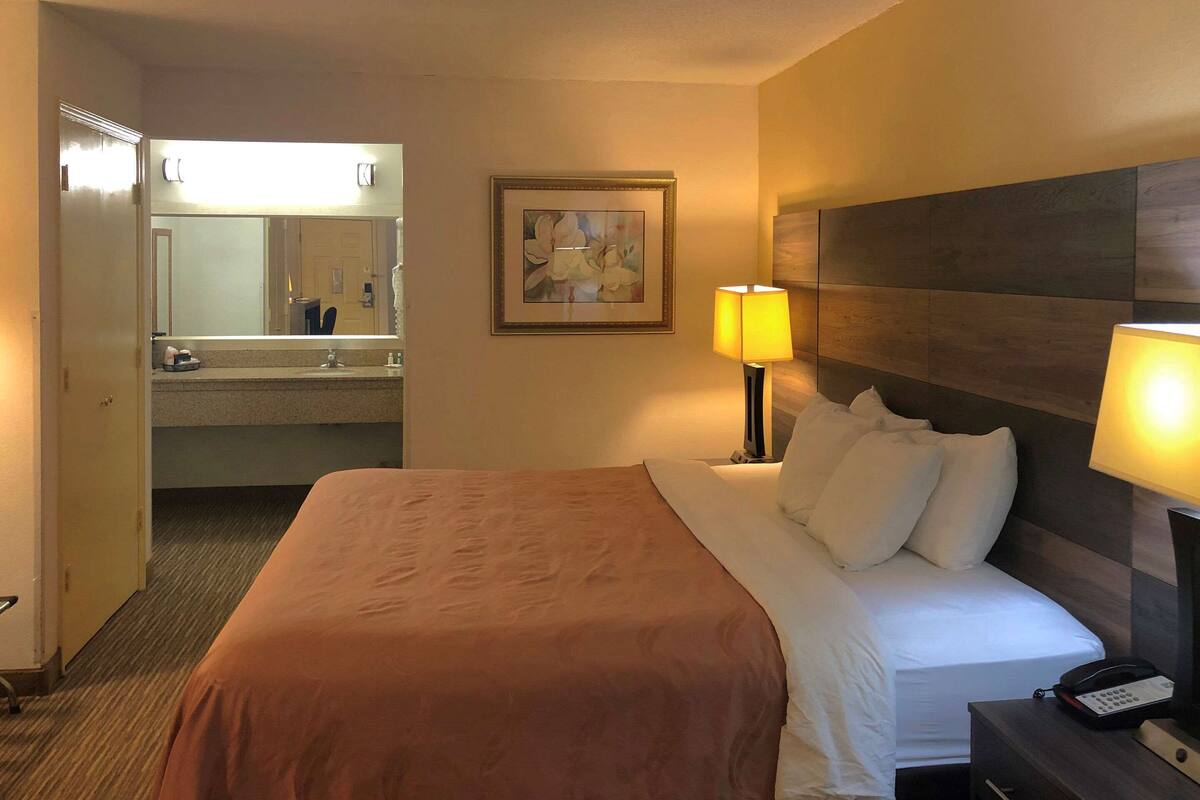 A hotel room with a bed, bedside lamps, a nightstand, a desk, a chair, a mirror, and a bathroom visible through an open door.