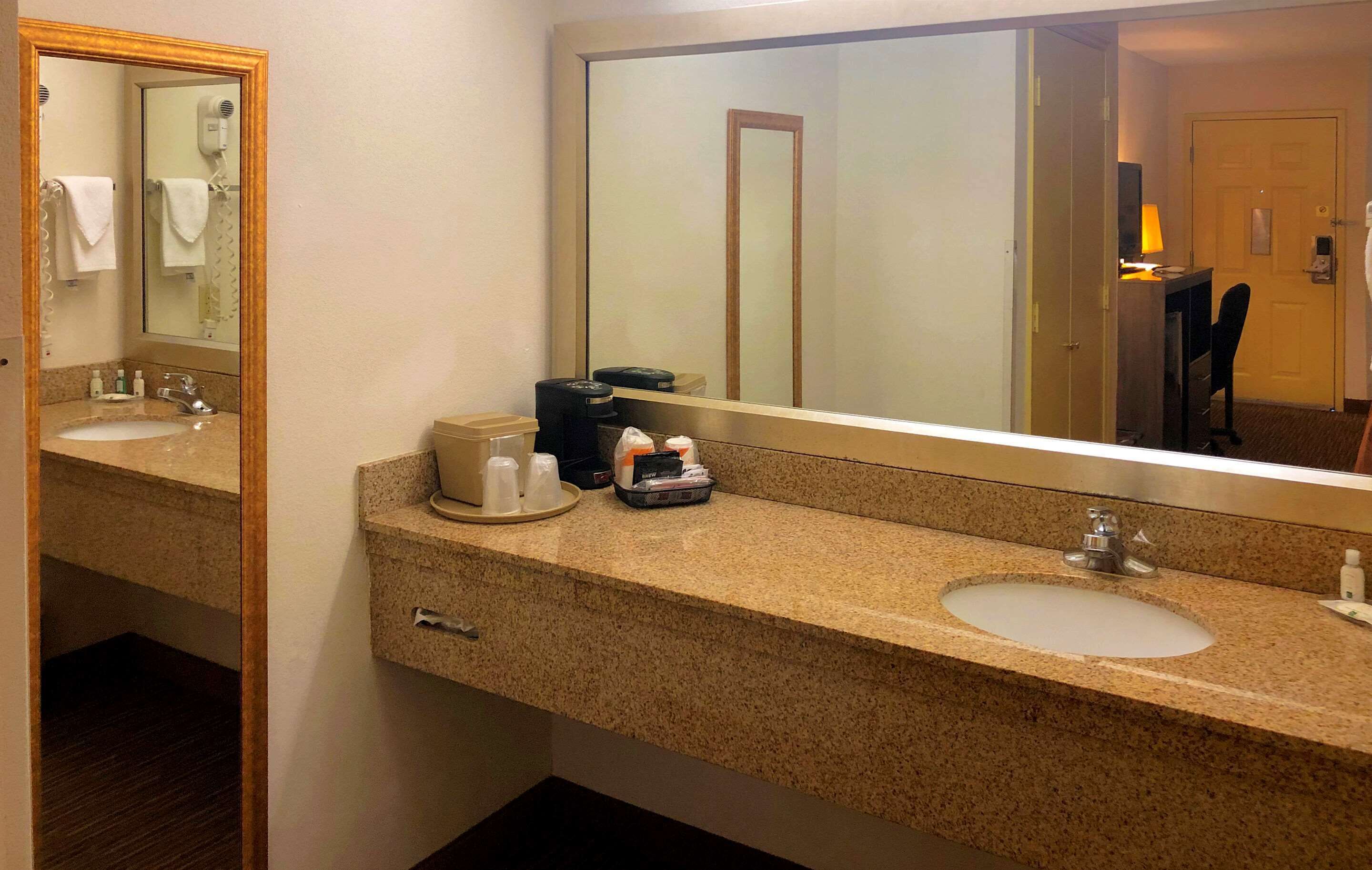 Un bagno d'hotel con due lavandini, uno specchio grande, un asciugacapelli e una macchina da caffè sul ripiano.