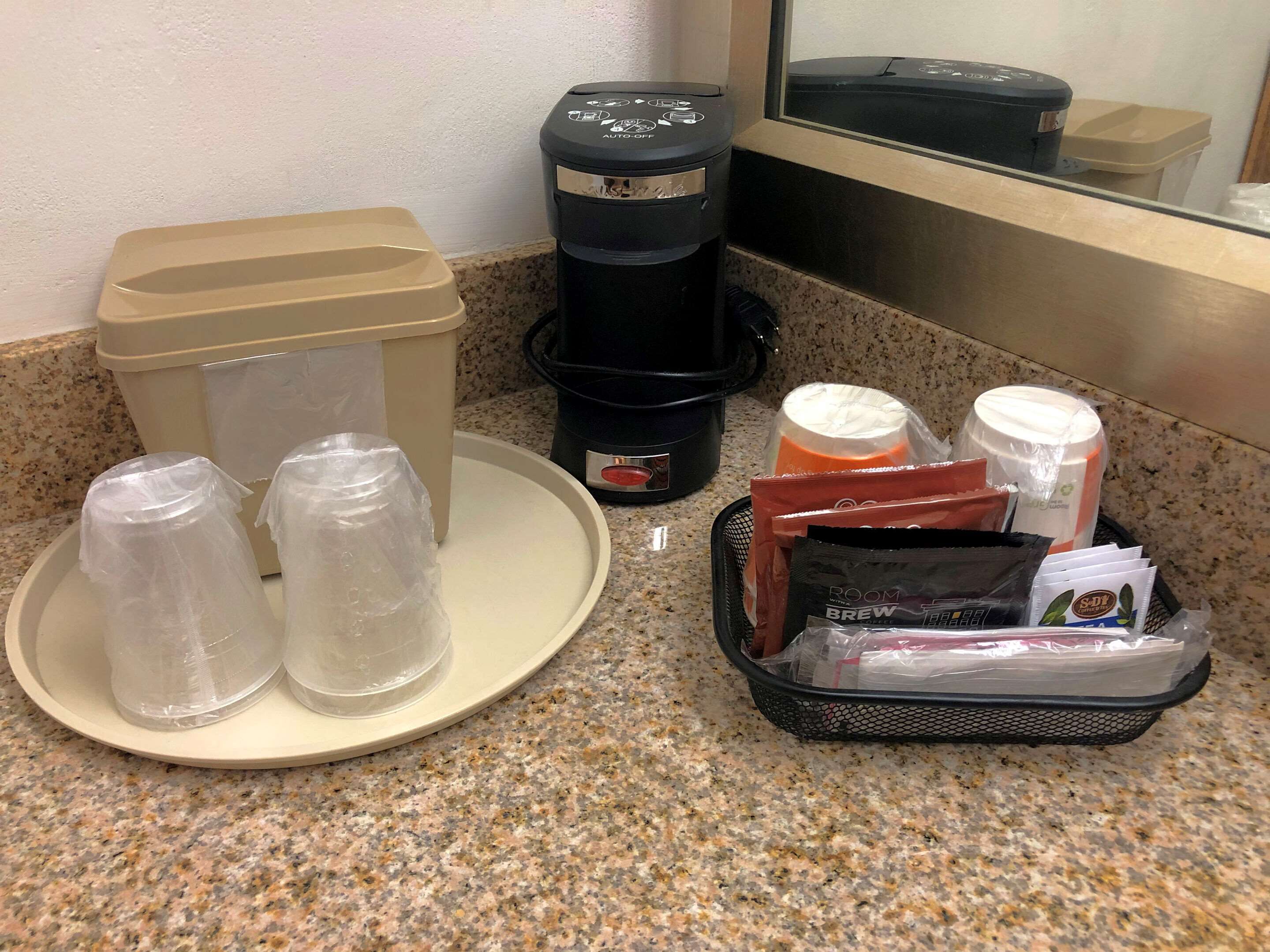 A kitchen counter with a coffee maker, a trash can, a plate with two plastic cups, and a basket containing coffee pods and a milk carton.