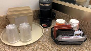 A kitchen counter with a coffee maker, a trash can, a plate with two plastic cups, and a basket containing coffee pods and a milk carton.