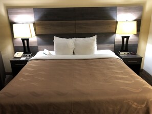 A hotel room with a large bed, two bedside tables, a nightstand, a lamp, a clock, and a phone.