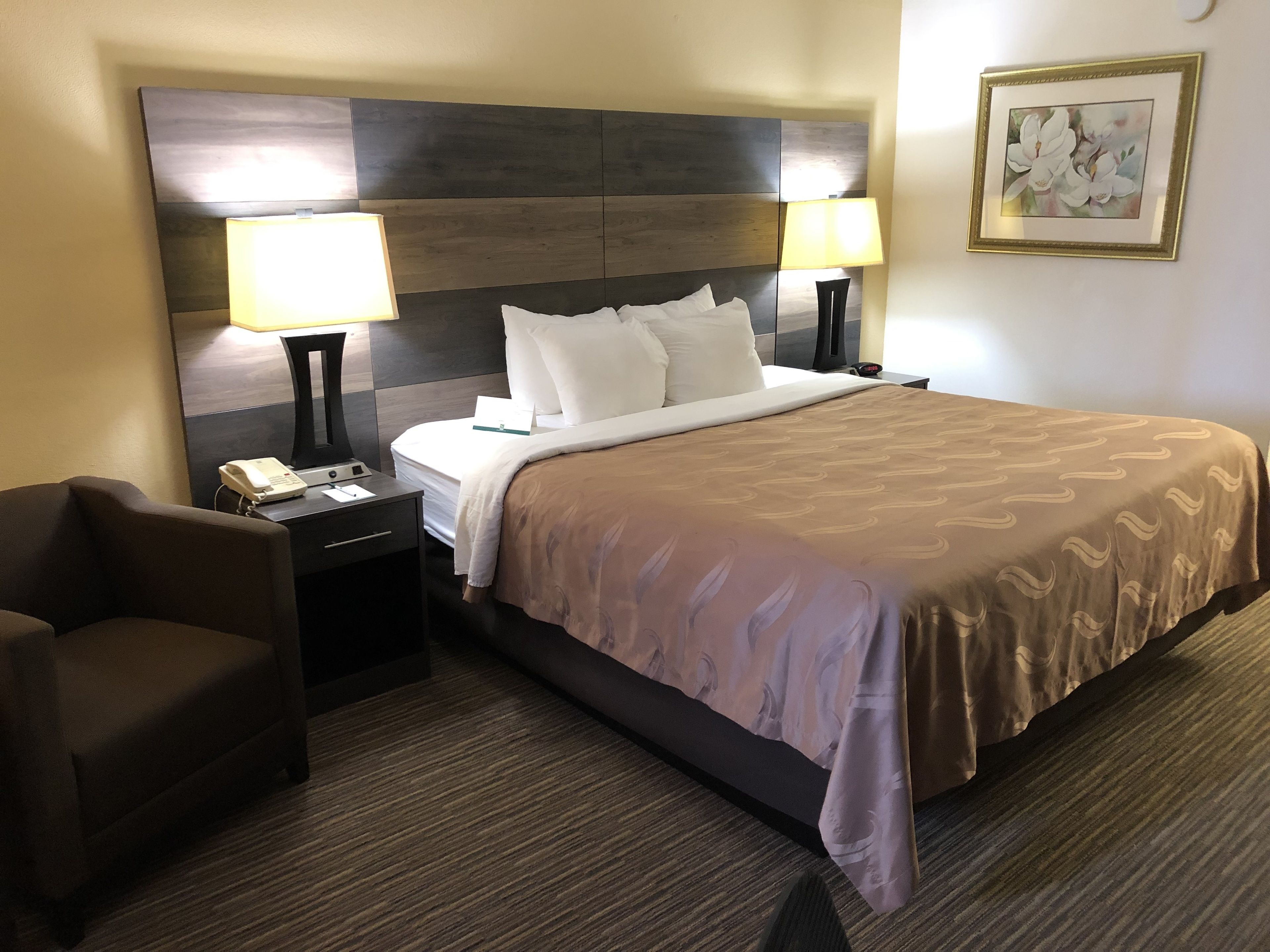 A hotel room with a large bed, two bedside lamps, a chair, a nightstand with a phone, and a framed picture on the wall.