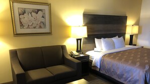 A hotel room with a sofa, a small table, a bed with a patterned bedspread, two bedside lamps, a nightstand with a telephone, and a framed floral artwork on the wall. - Quality Inn Bennettsville (Bennettsville)