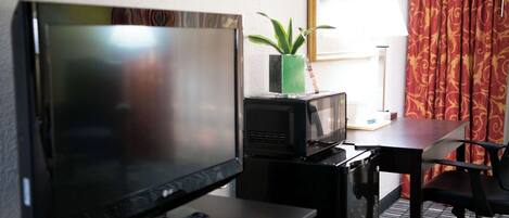 A hotel room with a TV, a microwave, a small table, a chair, a plant, and a window with curtains.