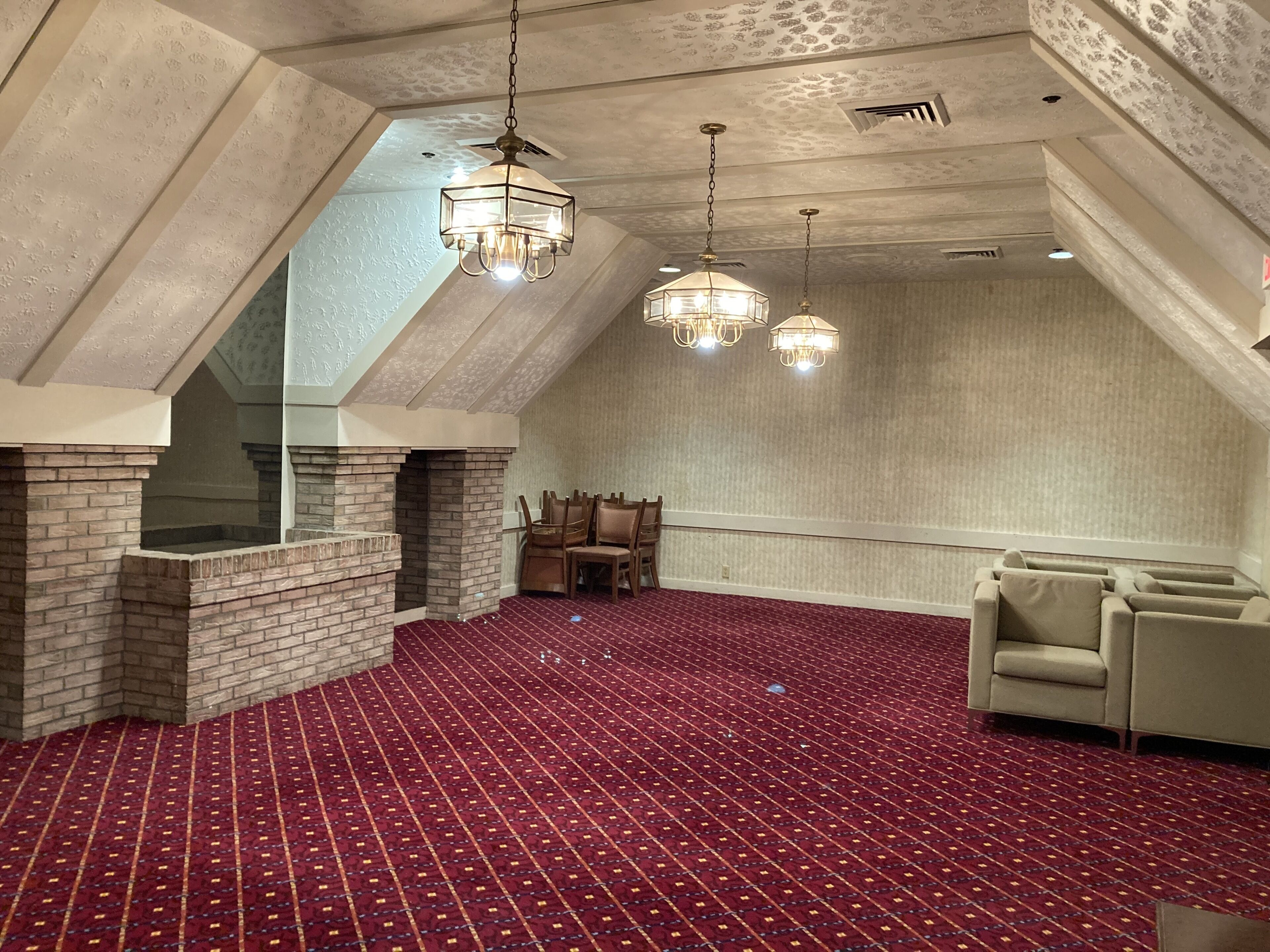 A spacious room with a high ceiling, a brick fireplace, patterned carpet, and two hanging chandeliers.