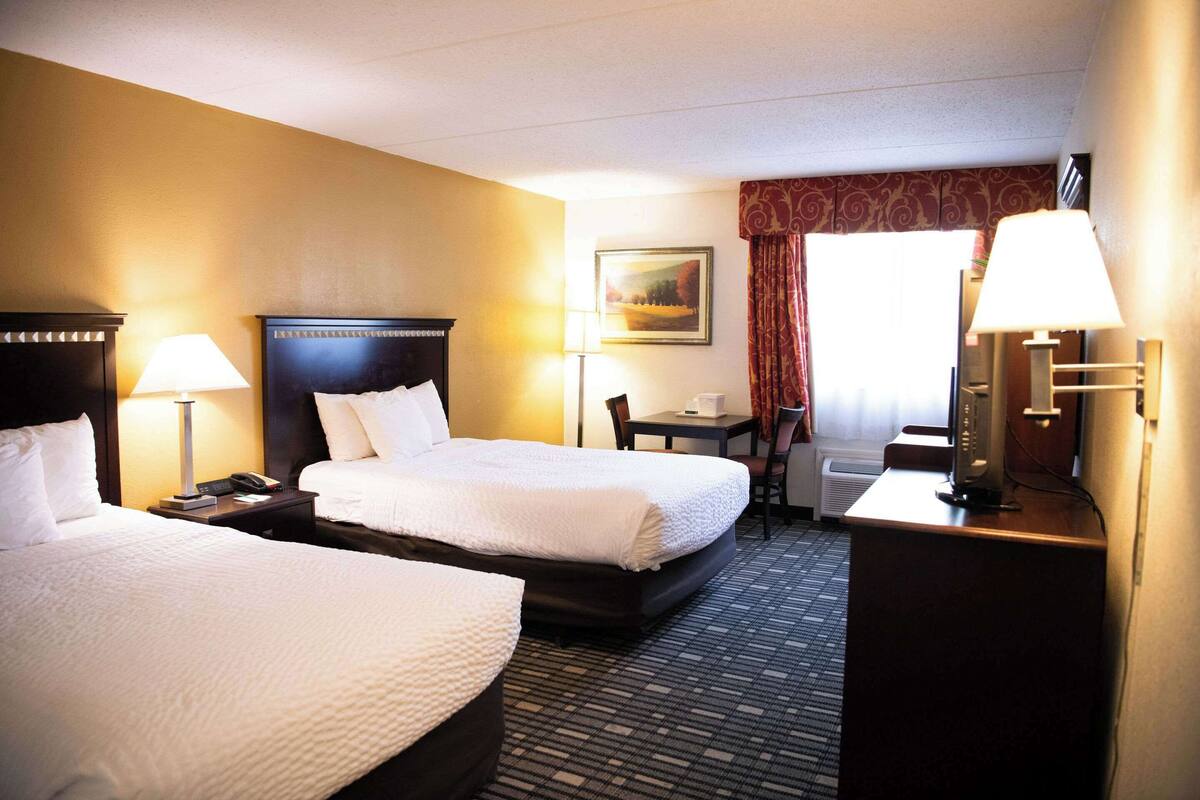 Quarto de hotel com duas camas, uma escrivaninha, uma cadeira, um abajur e um quadro na parede.