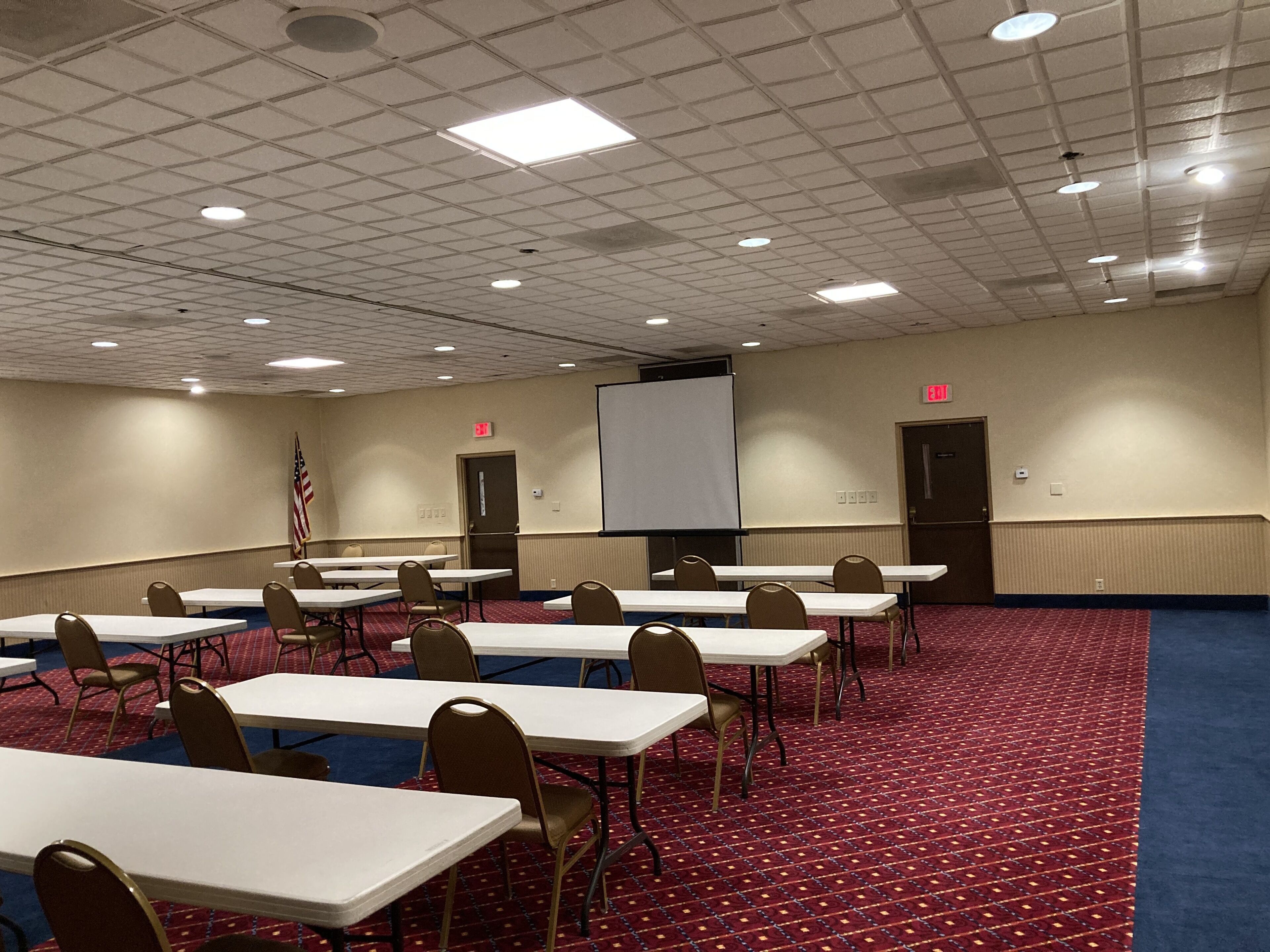 A conference room with tables and chairs, a projector screen, an American flag, and a carpeted floor.