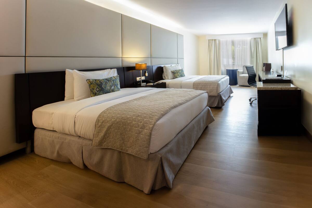 A hotel room with two beds, a desk, a chair, a flat-screen TV, and a large window with curtains.