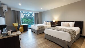 A hotel room with two beds, a desk with a coffee maker, a chair, and a view of trees through the window.