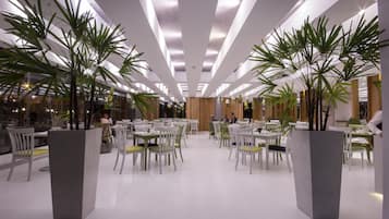 A modern dining area with white chairs and tables, large potted plants, and a wooden accent wall.