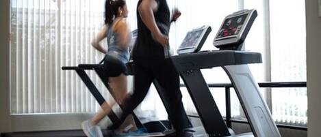 Two people running on a treadmill in a gym with a large window and a television screen.