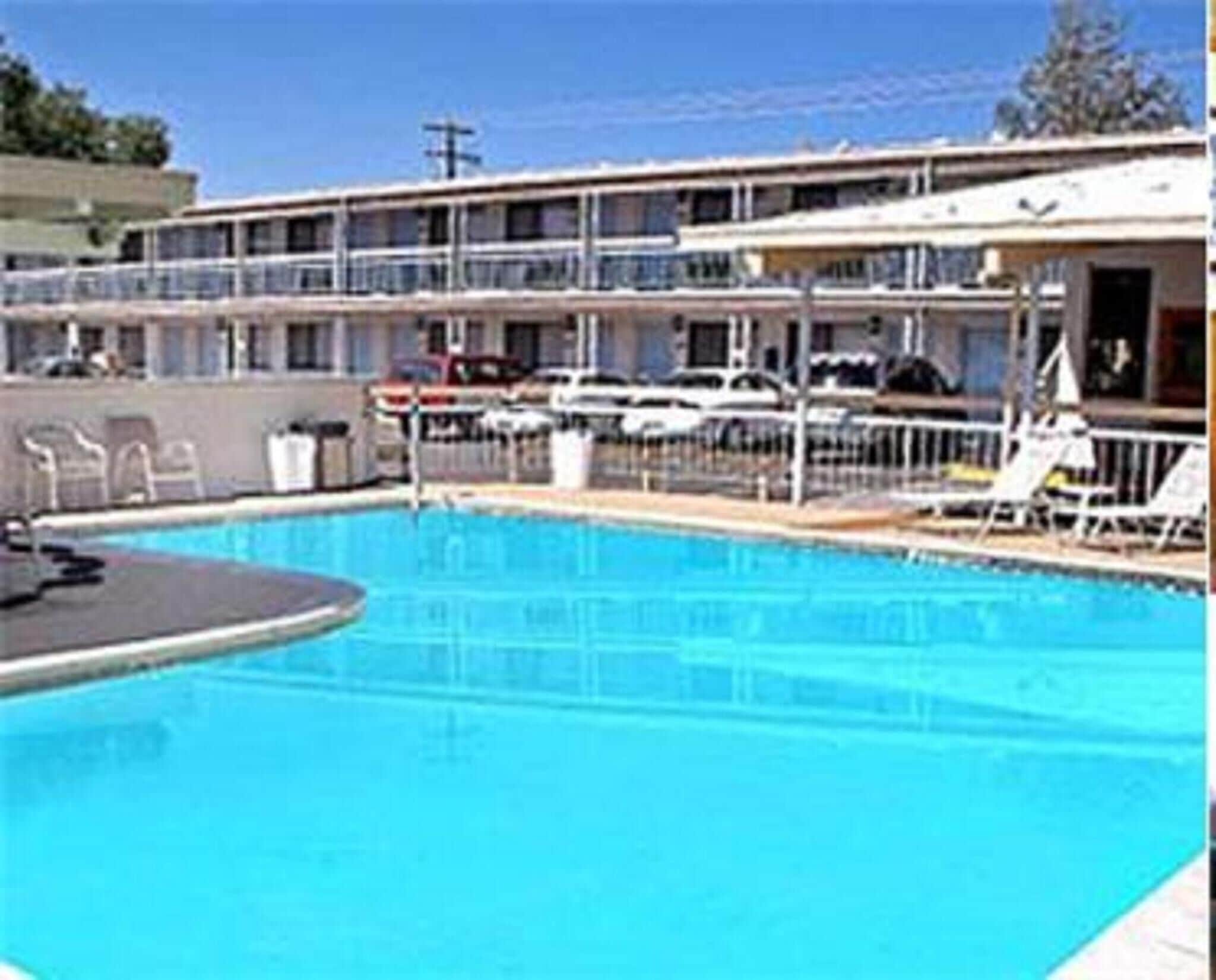 Piscina com espreguiçadeiras, área coberta e um edifício com varandas ao fundo.