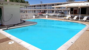 A rectangular swimming pool with a clear blue water, surrounded by a white pool deck. There are lounge chairs and a covered area with a roof. - Holiday Motel (Winnemucca)