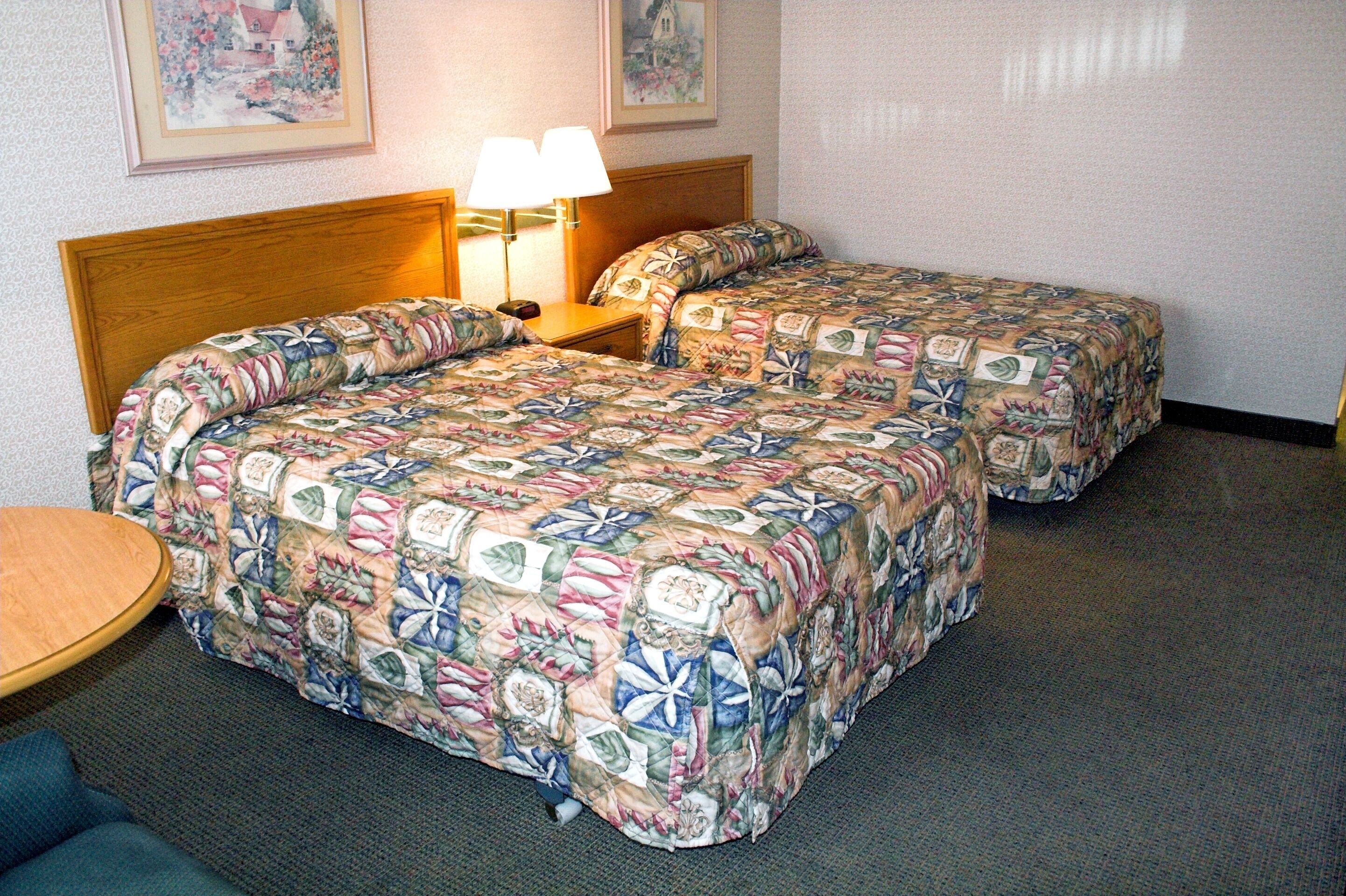A hotel room with two beds, each with a floral-patterned bedspread, a wooden headboard, a bedside table with a lamp, and a small round table.