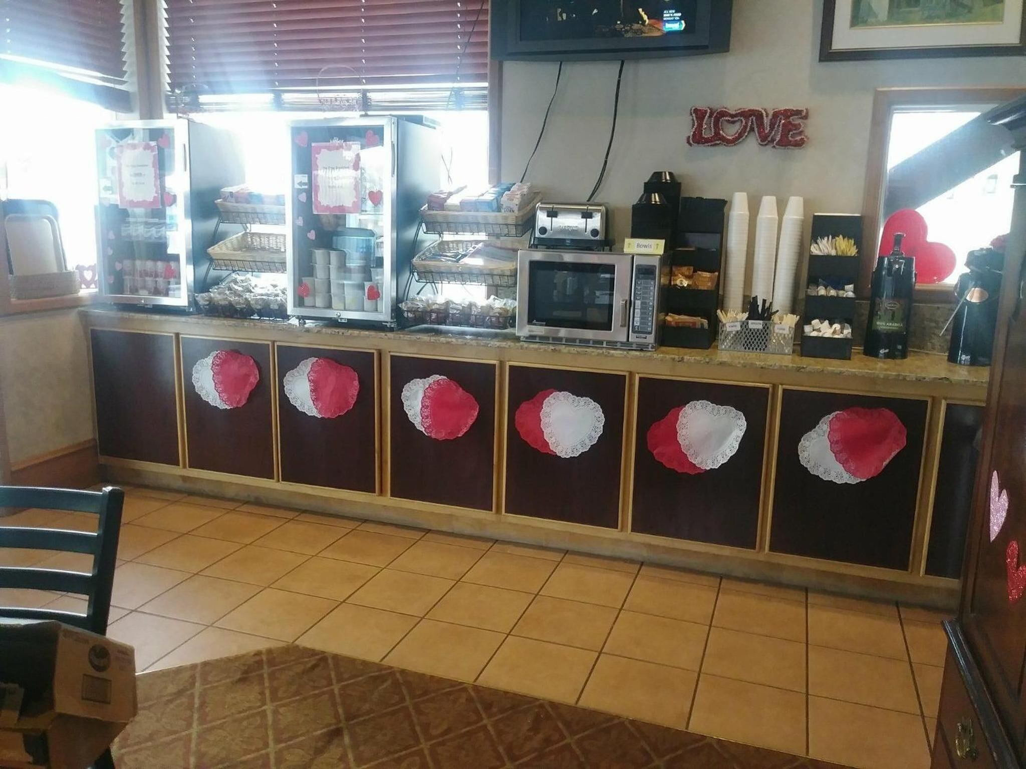 Un café con un mostrador que exhibe pasteles y equipo para café, un televisor montado en la pared y una decoración que dice 'LOVE'.