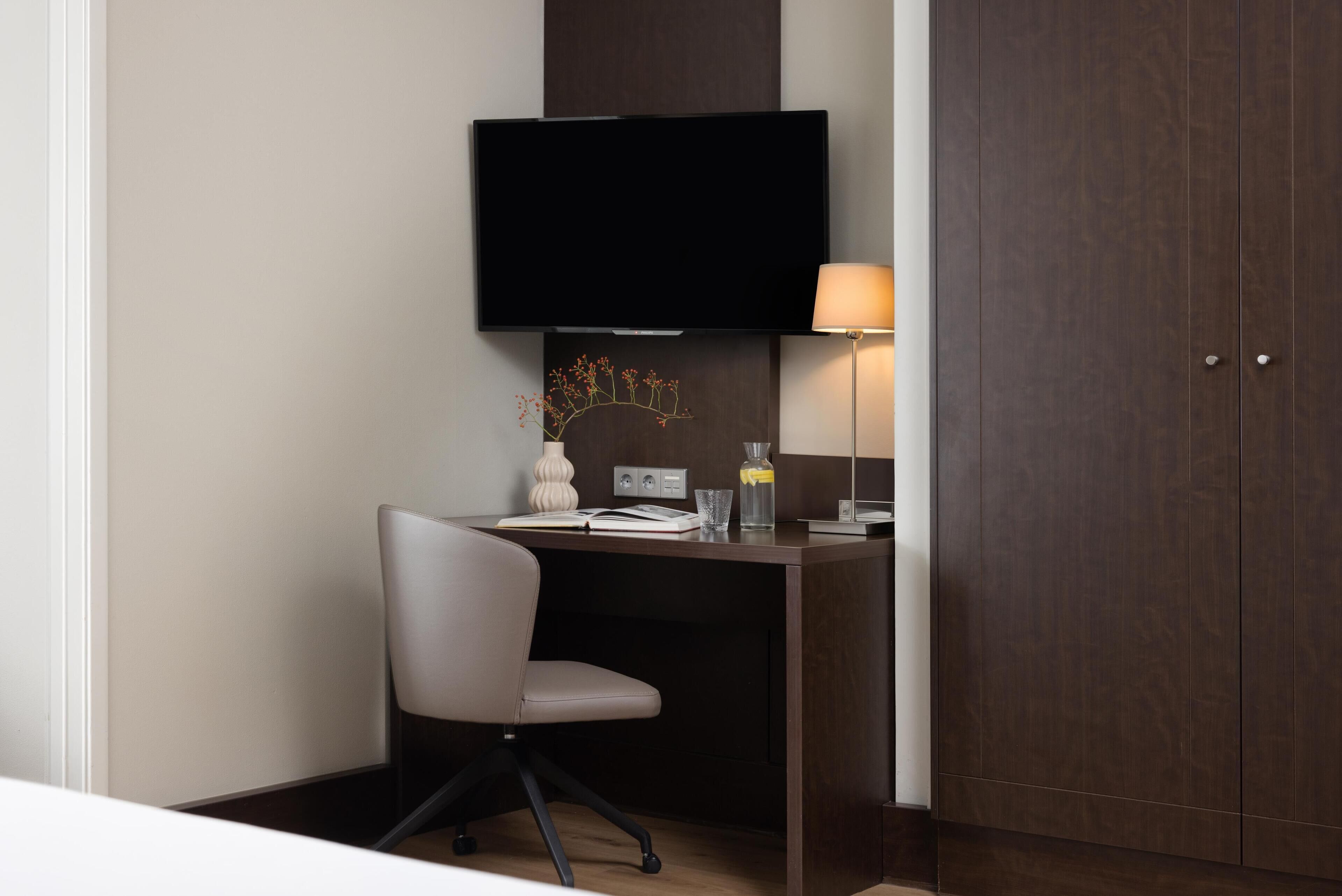 Een moderne hotelkamer met een bureau, stoel, flatscreen televisie, bedlampje en een kledingkast.