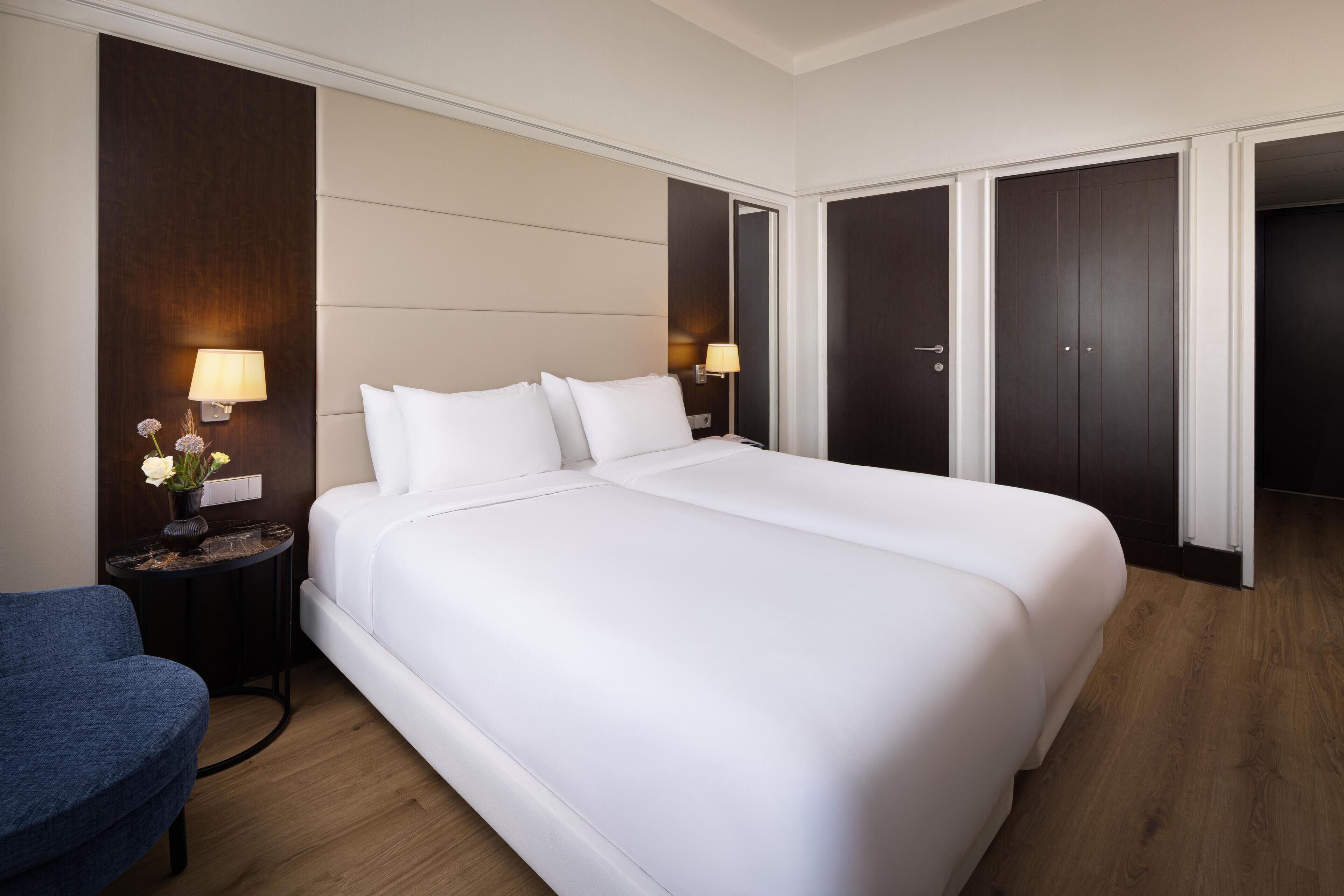 Quarto de hotel com uma cama grande, duas mesas de cabeceira com luminárias, uma mesa redonda pequena com um vaso de flores, uma cadeira azul e armários de madeira escura.