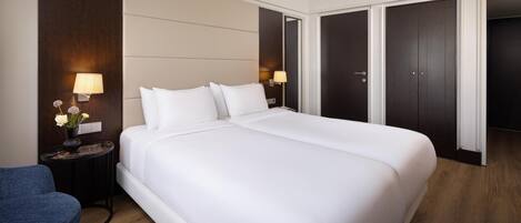 Una habitación de hotel con una cama grande, dos mesitas de noche con lámparas, una pequeña mesa redonda con un florero y una silla azul, además de armarios de madera oscura.