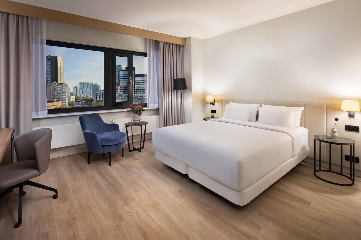 A hotel room with a large bed, a desk, a chair, a window with a city view, and two bedside lamps.